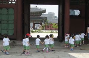 A kindergarten class on a fieldtrip files past Geunjeongmun. In the background, trees grow beside the once-buried stream. In the far background is scaffolding around Gwanghwamun's construction site.