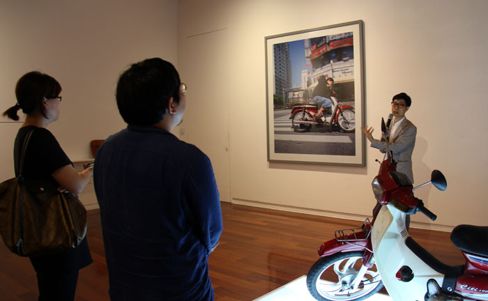 Visitors listen to an explanation from the exhibit organizer (right) about the framed photo of a delivery man on a scooter.