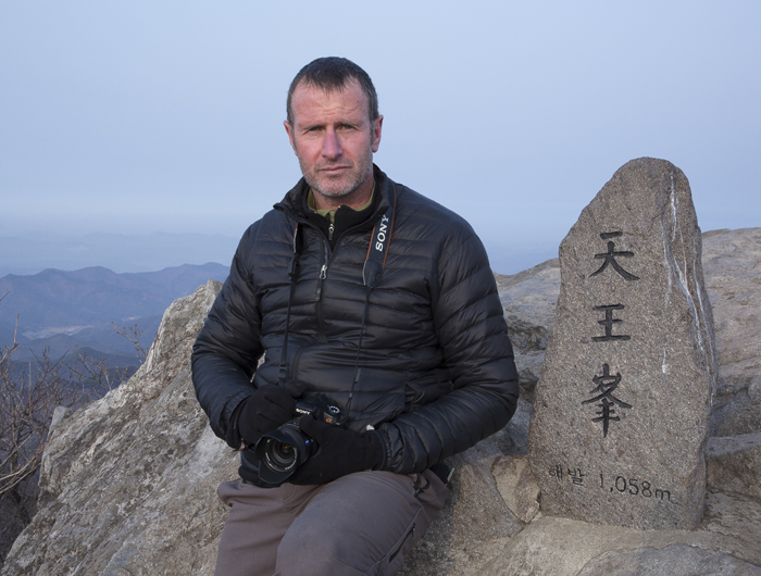 Roger Shepherd, armed with camera, sits atop the <i>Cheonwang-bong</i> Peak (천왕봉, 天王峰, King of Heaven Peak), the highest point on Songnisan Mountain (속리산, 俗離山, Apart From Society Mountain), in Chungcheongbuk-do (North Chungcheong Province). Other “King of Heaven Peaks” exist on other mountain tops. (photo: Roger Shepherd)