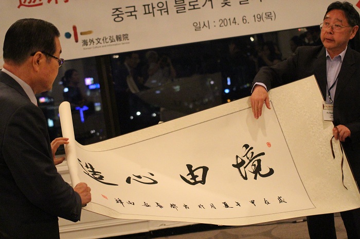 The head of the Chinese delegation, Wu Changsheng, presents to Lee Se-ki, head of the Korea-China Friendship Association, a gigantic paper with a famous Chinese phrase written on it. It reads <i>Jing You Xin Zao</i>, or 境 由 心 造), meaning that, 