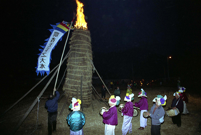 People perform some <i>nongak</i> farmers’ music in 1993 in front of a large <i>daljip</i> bonfire (달집) on the night of Jeongwol Daeboreum, the first full moon of the lunar year. A large flag hanging on the bonfire says that, ‘Farmers are the great foundation of the world.' In a traditional, agrarian Korea, people lit the straw structure on fire to remove any misfortune and to wish for a good harvest.