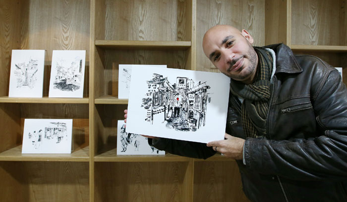 Dahmani poses for a photo while holding one of his works of art. A man wearing a red mask shows up in many of the illustrations.