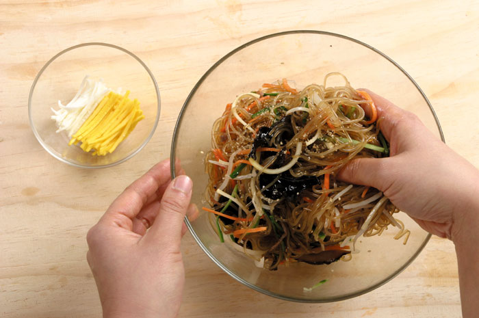 Pork, spinach, oyster mushrooms and other ingredients may be added to <i>japchae</i>. It's served with the noodles mixed with the other ingredients and topped with yellow and white egg strips.
