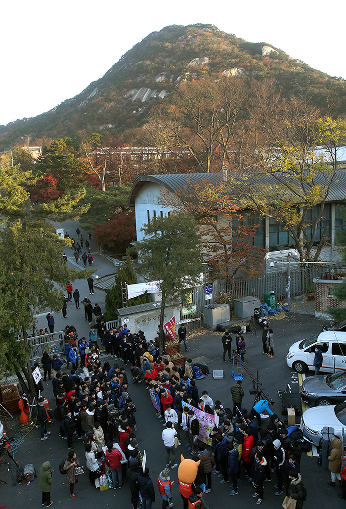 Even before 6:00 a.m., the front gate of Kyungbok High School is crowded with students, parents and teachers who headed out in below freezing temperatures to cheer on the test-takers. Reporters nearby cover the exam-day story.
