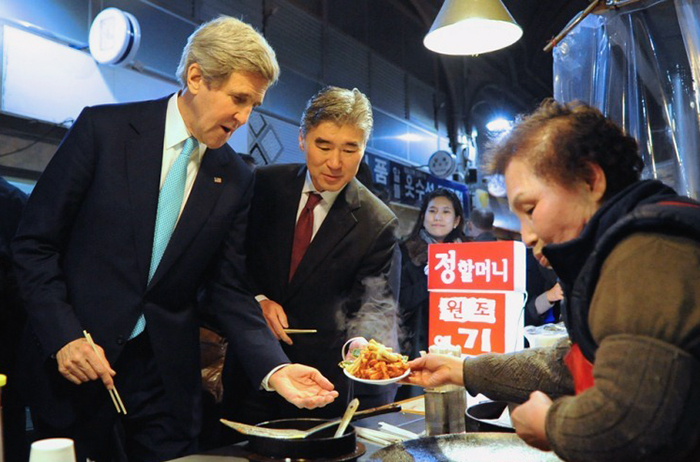 U.S. Secretary of State John Kerry (left) buys a dish of oil-fried <i>tteokbokki</i> at Tongin Market on February 13. U.S. Ambassador to Korea Sung Kim announced in the official online community of the U.S. embassy in Seoul that he’d included a visit to the traditional market in the secretary of state’s itinerary so that Kerry could feel the real Korea, meet real people and enjoy a specialty of the market. (image from the official online community of the U.S. embassy in Seoul)
