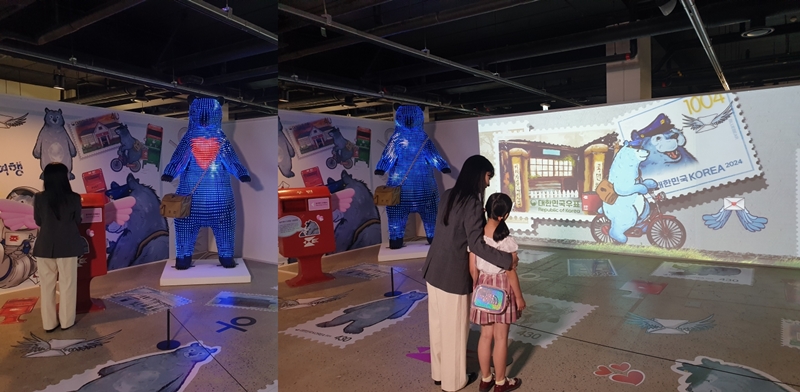 Visitors to the 2024 Korea Stamp Exhibition on its opening day of July 18 look at a work by artist Lee Jae-hyung featuring a blue bear traveling through the 140 years of domestic stamps on Nodeulseom Island in Seoul. (Koh Hyunjeong)
