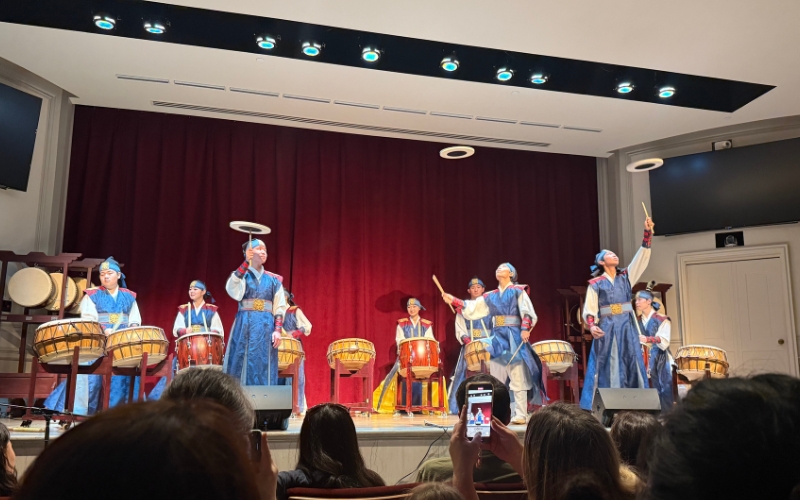 Korean drummers performing with skill and energy, as three young participants expertly toss discs into the air in front of the audience.