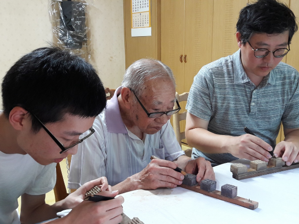Fifth generation Master Kim Gi-ho, fourth generation Grand Master Kim Deok-hwan and sixth generation trainee Kim Jinho(from right) work on carving wooden printing block for gold leaf imprinting at their workshop in Buckchon Hanok Village, Seoul Korea on August 2016, demonstrating the transmission of the craft across three generation together. Picture credit- Kim Jinho