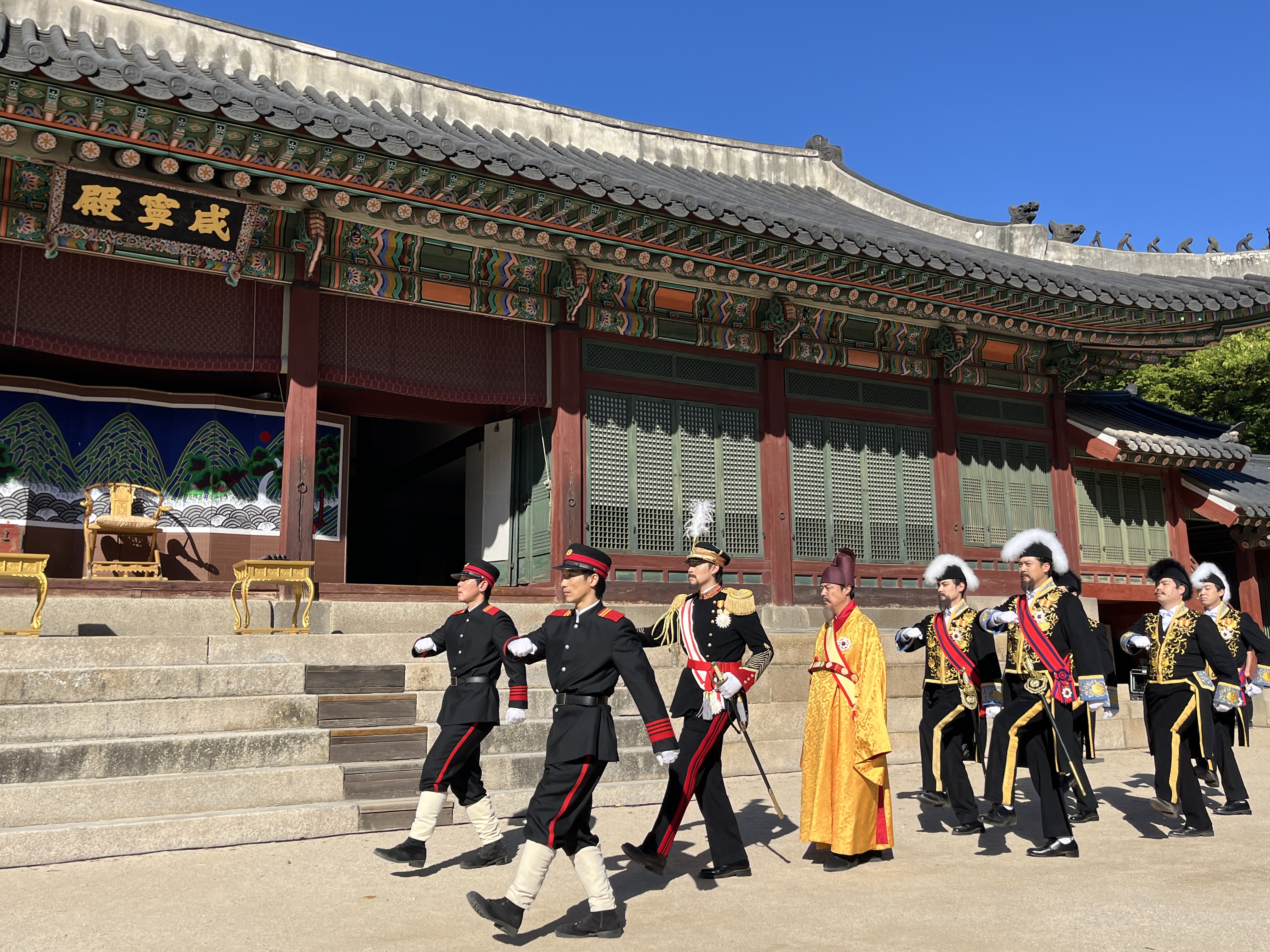 An actor portraying Emperor Gojong (fourth from left) enters Hamnyeongjeon Hall accompanied by officials and soldiers, moving slowly to the rhythm of the gentle background music.