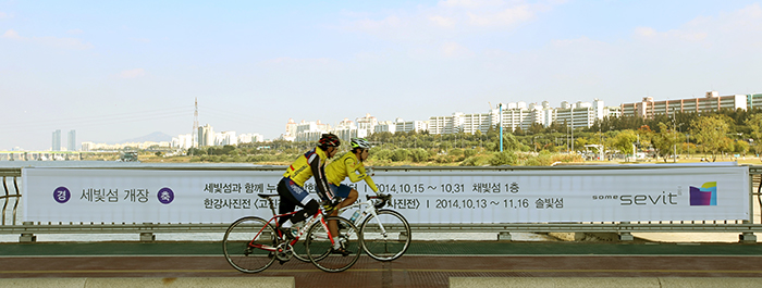 Two bicyclists ride along the Jamsu Bridge, the lower level of the Banpo Bridge, on October 15, as they pass a banner advertising the opening of the Some Sevit floating buildings. 