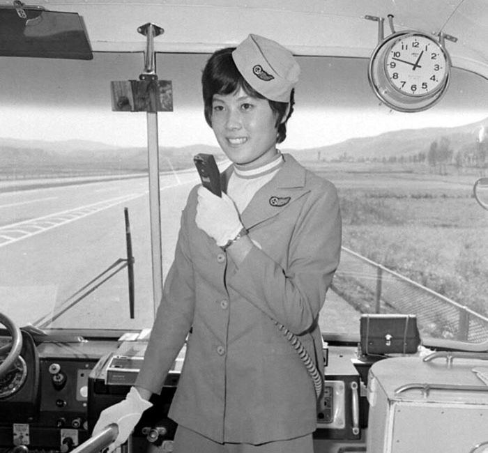 In the past, a bus attendant, usually a woman, was on board to collect the fares and to assist the bus driver. This photo shows a bus attendant wearing a uniform and holding a microphone while working on board an express bus in 1973.