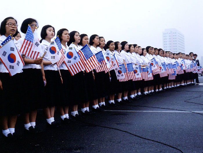 Students gather to greet U.S. President Jimmy Carter on his visit to Korea in 1979. The students are all in uniform and are holding the flags of both Korea and the U.S.