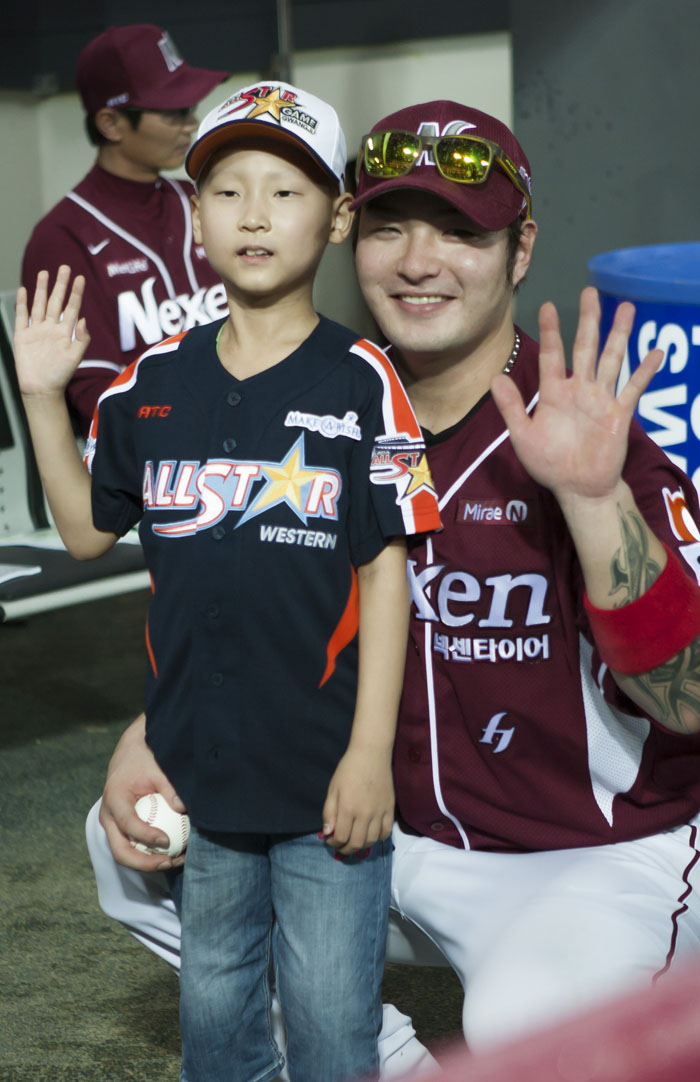 Park Ju-sang (left), an eight-year-old boy suffering from leukemia, waves alongside Park Byung-ho, first baseman for the Nexen Heroes. After the bottom of the fifth inning at the 2014 Professional Baseball All Star game, Ju-sang stood in the batter's box, swung as hard as he could and scored a run, advancing to first base. (photo courtesy of Make-A-Wish Korea) 