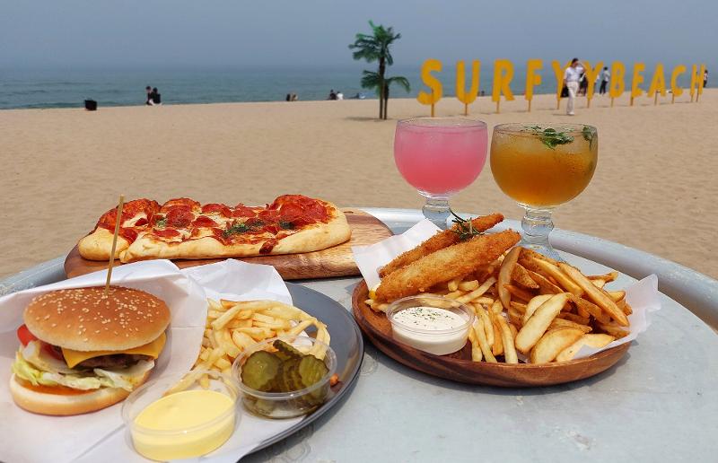 A slew of foods like pizza, hamburgers, and fish and chips as well as beer and cocktails are offered at the sunset bars of Surfyy Beach, creating an exotic experience for visitors. (Anais Faure)